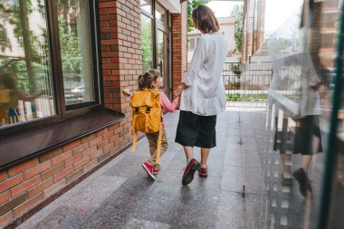Mother leading her daughter at school. Back view of young woman and little 6 years old girl near elementary school building. Back to school concept.