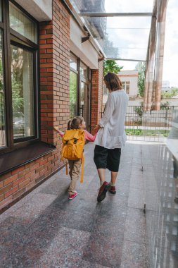 Mother leading her daughter at school. Back view of young woman and little 6 years old girl near elementary school building. Back to school concept.