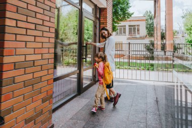 Mother leading her daughter at school. Young woman and little 6 years old girl near elementary school building opening the door. Back to school concept.