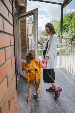 Mother leading her daughter at school. Young woman and little 6 years old girl near elementary school building opening the door. Back to school concept.