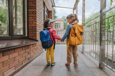 Back view of two little girls with backpacks holding hands going to an elementary school. Back to school concept. Selective focus on girls.