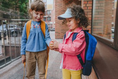 Back view of two little girls with backpacks going to an elementary school. Friend giving an apple. Back to school concept. Selective focus on girls.