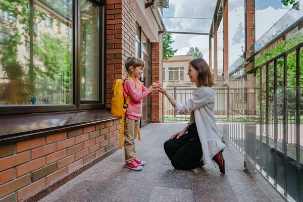 Mother leading her daughter at school giving an apple. Young woman and little 6 years old girl near elementary school building. Back to school concept.