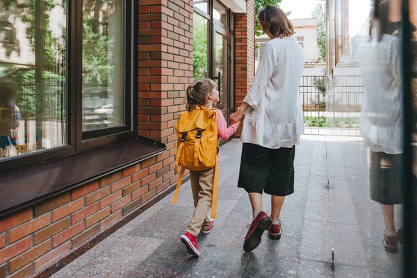 Mother leading her daughter at school. Back view of young woman and little 6 years old girl near elementary school building. Back to school concept.