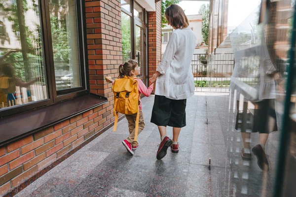 Mother leading her daughter at school. Back view of young woman and little 6 years old girl near elementary school building. Back to school concept.