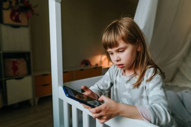 Little girl using smartphone sitting on the bed in handmade house. Generation alpha using technology. Social media concept.