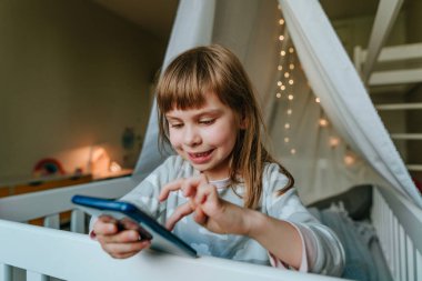 Little girl using smartphone sitting on the bed in handmade house. Generation alpha using technology. Social media concept.