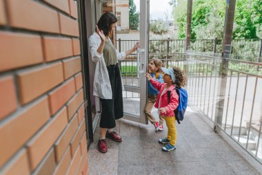Happy smiling woman teacher greeting elementary school students at first day at school standing near the open door outside. Selective focus.