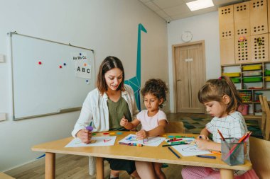 English lesson at elementary school or kindergarten. Students learning alphabet and colors with teacher coloring the letter A sitting in the classroom. Selective focus.