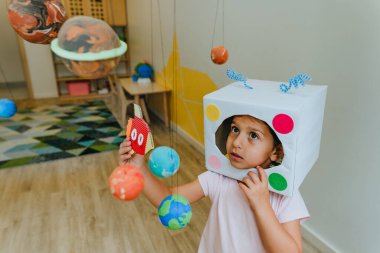 Funny little girl wearing handmade helmet playing with paper spaceship learning Solar system planets models at home or kindergarten. Education science concept. Selective focus.