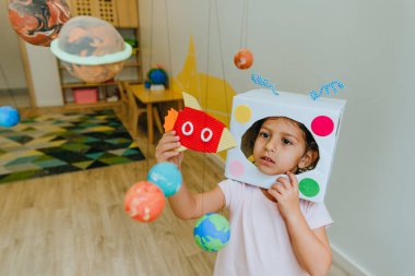 Funny little girl wearing handmade helmet playing with paper spaceship learning Solar system planets models at home or kindergarten. Education science concept. Selective focus.