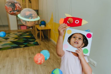 Funny little girl wearing handmade helmet playing with paper spaceship learning Solar system planets models at home or kindergarten. Education science concept. Selective focus.