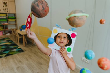 Funny little girl wearing handmade helmet playing with paper spaceship learning Solar system planets models at home or kindergarten. Education science concept. Selective focus.