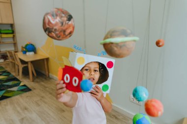 Funny little girl wearing handmade helmet playing with paper spaceship learning Solar system planets models at home or kindergarten. Education science concept. Selective focus.