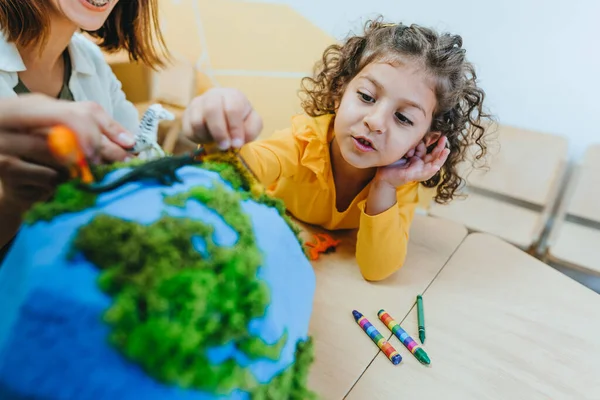 Natural science or Geography lesson at elementary school or kindergarten. Students playing with wild animals toys on handmade globe in the classroom. Selective focus.