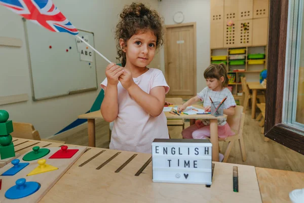 English language lesson at elementary school or kindergarten. Little girl holding UK Union Jack flag standing near inscription English time in the classroom. Selective focus on the girl.