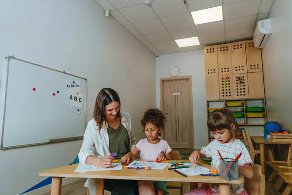 English lesson at elementary school or kindergarten. Students learning alphabet and colors with teacher coloring the letter A sitting in the classroom. Selective focus.