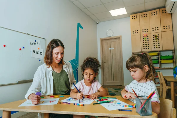 English lesson at elementary school or kindergarten. Students learning alphabet and colors with teacher coloring the letter A sitting in the classroom. Selective focus.