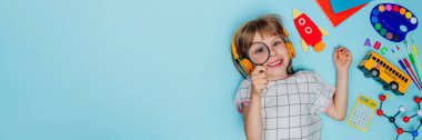 Etrafı mavi arka planda duran farklı okul kırtasiyeleriyle çevrili büyüteçle çevrili komik kız. Üst manzara, düz yatış. Okul konseptine geri dönelim. Panorama. Kopyalama alanı olan yatay pankart.