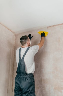 Tulum, eldiven ve koruyucu yüz maskesi takan, plastik spatula kullanarak duvara fiberglas yüzey dokusu uygulayan erkek işçi, sıva ve boyama için yüzey hazırlığı. Seçici odak.