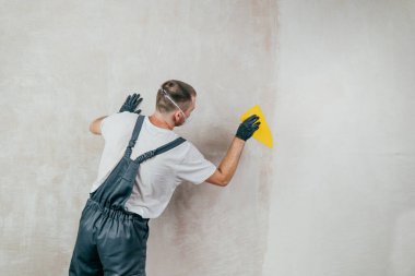 Tulum, eldiven ve koruyucu yüz maskesi takan, plastik spatula kullanarak duvara fiberglas yüzey dokusu uygulayan erkek işçi, sıva ve boyama için yüzey hazırlığı. Seçici odak.