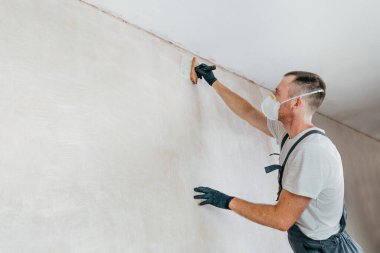 Tulumlu, eldivenli ve koruyucu maskeli duvarları fırçayla yapıştıran bir erkek işçi. Seçici odaklanma. Metin için boş boşluk.