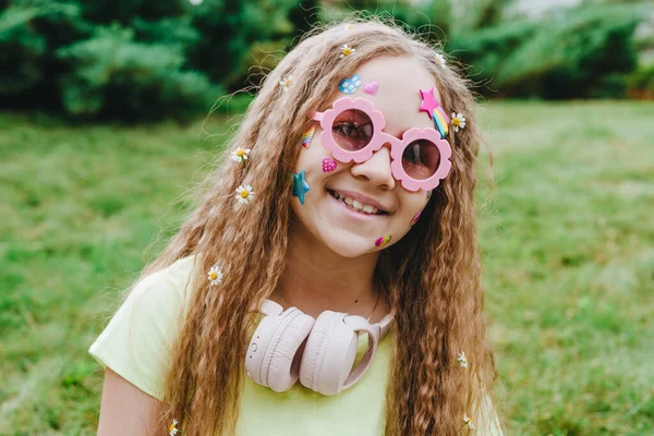 可爱的少女 满脸贴纸 戴着粉色眼镜 头戴耳机 站在绿草的背景上 — 图库照片