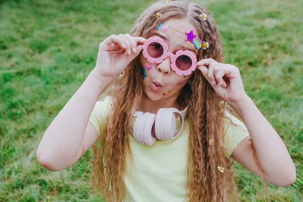 可愛いですティーン女の子とともにステッカー彼女の顔身に着けていますピンクガラスとヘッドフォンとともにカモミール花で巻き毛でポーズ緑の草の背景 — ストック写真