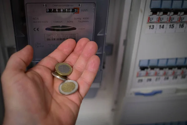 in one hand are several euro coins in front of a three-phase meter