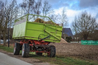 Yeşil bir traktör römorku yol kenarında bir tarlanın yanında duruyor.