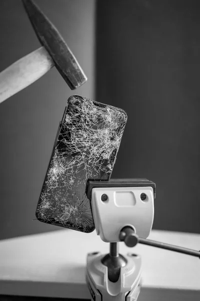 a completely destroyed cell phone is clamped in a vice and above it is a hammer