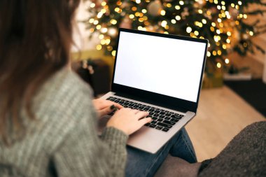 Kadın dizüstü bilgisayarda çalışıyor. Boş ekran ve taklit alanı var. Noel kutlaması için Noel ağacı ve çelenk ışıklarıyla dekore edilmiş ev.