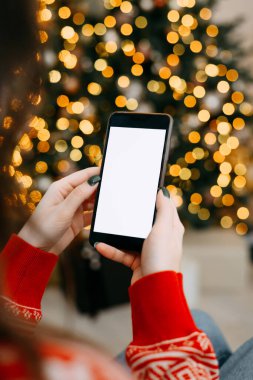 Mesaj atmak için yeri olan bir Noel modeli. Noel reklamı, uygulama şablonu. Işıkları olan altın güzel Noel ağacının arka planında boş ekranlı telefonu tutan eller..