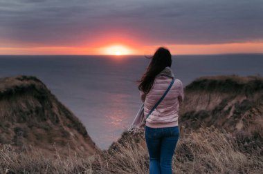 Gün batımında doğayı düşünen bir kadının arka plan görüntüsü. Gezgin yürüyüşçü deniz manzarasının tadını çıkarıyor..