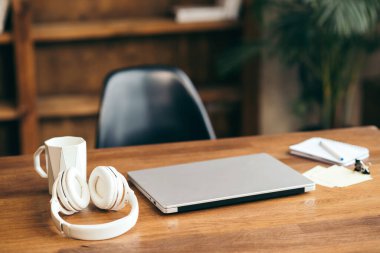 İş yeri konsepti. Bardaklı, kulaklıklı gümüş bir dizüstü bilgisayar ve ev içinde bir ahşap masa üzerinde defter. Serbest ofis.