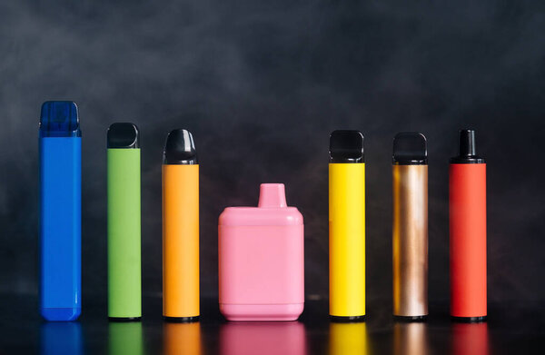 A set of colorful disposable electronic cigarettes of different shapes on a black background with smoke. 