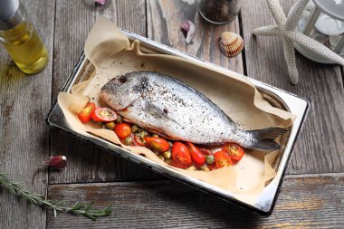 Tüm deniz çipurası sebzelerle dolu, ahşap bir masada, fırın kabına baharat serpiştirilmiş. Parşömen kağıdıyla kaplanmış bir tavada balık pişirmek.. 