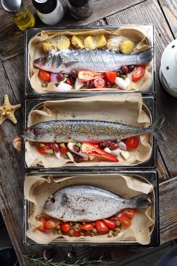 Tüm levrek, uskumru ve çipura, ahşap bir masada, fırın tabaklarına baharat serpiştirilmiş. Parşömen kağıdıyla kaplanmış bir tavada sebzeli balık pişirmek.. 