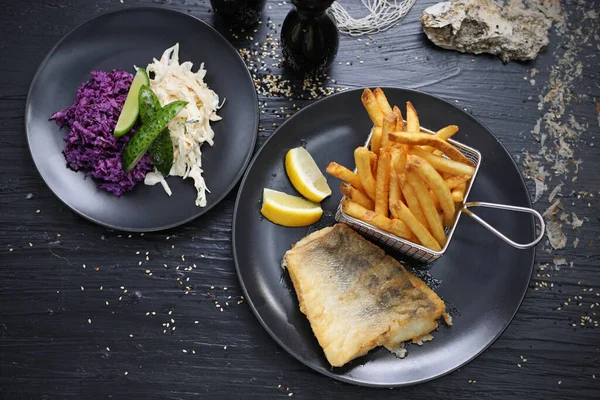 Fried fish fillets served with potato fries in a metal serving basket and salad mix, on black plates, top view. Composition with a fish and chips lunch set in a fish restaurant.