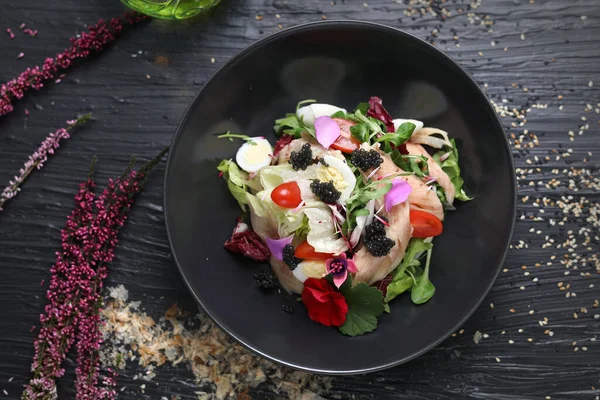 Salad with vegetables, fresh fish, caviar and quail egg, decorated with edible flowers. Fish salad in a black bowl, top view.