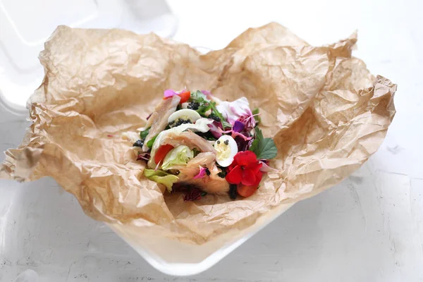 Salad with vegetables, fresh fish, caviar and quail egg, decorated with edible flowers. Fish salad in a takeaway box.