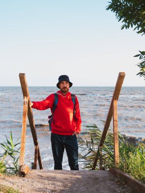Sırt çantalı, güz mevsimi arka planında kuru sazlıklar olan kırmızı kapüşonlu sakallı bir adam. Otantik erkek turist yaşam tarzı fotoğrafı. Hippi adam dışarıda. Solo seyahat macera konsepti aktif yürüyüş sırt çantası