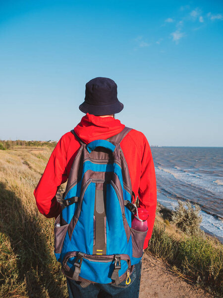 Тысячелетний человек в ковшовой шляпе красный рюкзак с капюшоном на осеннем фоне моря. Фотография подлинного мужского туристического образа жизни. Хипстерский парень под открытым небом