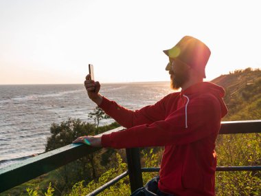 Sakallı milenyum adamı, kırmızı kapüşonlu sırt çantasıyla bankta akıllı telefonuyla sonbahar deniz manzarasının fotoğrafını çekiyor. Otantik erkek turist blogcu adam Solo seyahat macerası. Günlük gezintisi sırt çantası.