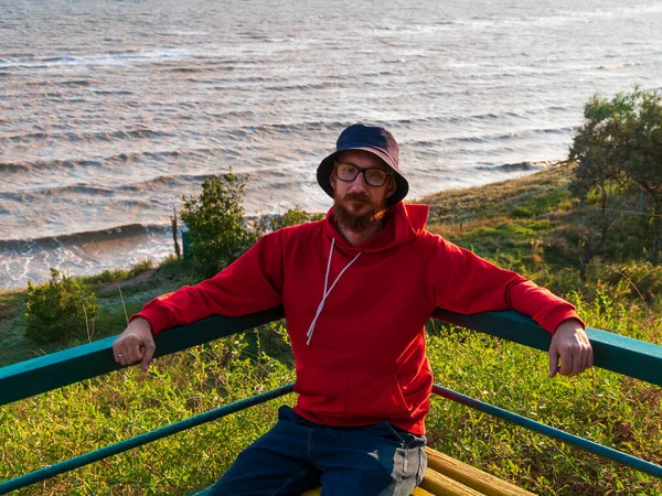 Millennial man in bucket hat red hoody eyeglasses on a bench with autumn sea view. Authentic male tourist lifestyle photo. Hipster guy outdoor. Solo travel adventure concept Active walking backpacking