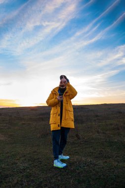 Sarı ceketli, akıllı telefonlu bir gezgin gün batımına bakarak tepede yürüyor. Cihaz yaşam tarzı olan otantik bir erkek turist. Açık havada gezen hippi adam Solo seyahat macerası. Aktif yürüyüş sırt çantası.