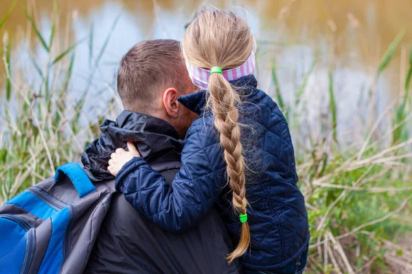 Baba ve kızı göl kenarında oturmuş doğa manzarasıyla sarılıyorlar. Saçları örgülü küçük sarışın kız babasıyla dışarıda yürüyor. Sevgi dolu çocuk, babayı kucakla. Babalar Günü, özgün aile, aşk yürüyüşü.