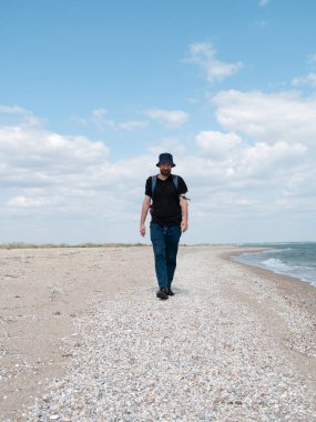 Kova şapkalı, siyah tişörtlü, boş sahilde yürüyen gülümseyen sakallı bin yıllık adam. Otantik erkek turist yaşam tarzı fotoğrafı. Hippi adam dışarıda. Solo seyahat macera konsepti aktif yürüyüş sırt çantası