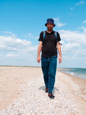 Kova şapkalı, siyah tişörtlü, boş sahilde yürüyen gülümseyen sakallı bin yıllık adam. Otantik erkek turist yaşam tarzı fotoğrafı. Hippi adam dışarıda. Solo seyahat macera konsepti aktif yürüyüş sırt çantası