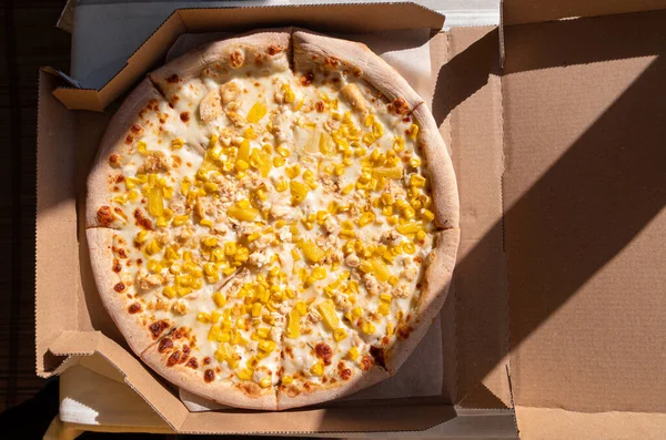 Tasty pizza with pineapples, corn and mozzarella cheese and white sauce in cardboard box on a table in daylight harsh shadows. Fresh baked creamy Hawaiian pizza crust italian cuisine delivery flat lay